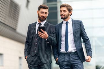 Young businessman in suit using mobile phone and showing something to his colleague while they standing near the modern building outdoors