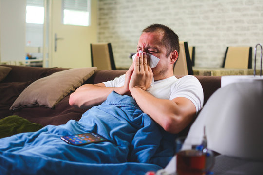 Sick man with headache sitting under the blanket. Sick man with seasonal infections, flu, allergy lying in bed. Sick male covered with a blanket lying in bed with high fever and a flu, resting.