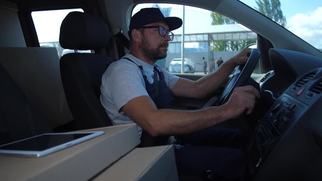 Serious delivery man in uniform setting route to client using navigator app while sitting in van. Adult worker of delivery service going to deliver order and using mobile phone