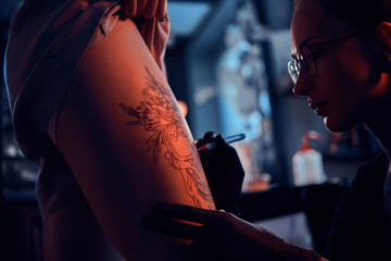 Attractive focused tattoo artist is creating new tattoo on young woman's leg at dark tattoo studio.