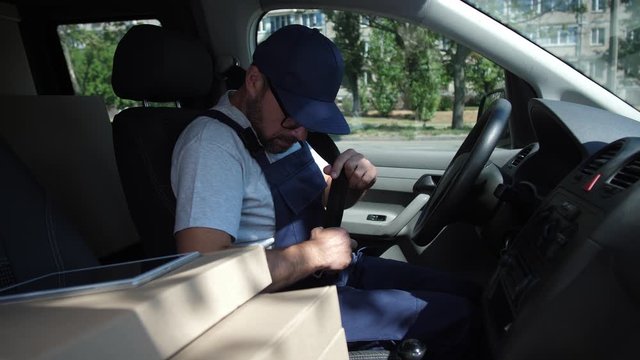 Adult worker of speed delivery service getting in car, fastening safety belt, starting and driving to deliver order. Delivery man in cap and glasses driving van, boxes and tablet pc lying next to him
