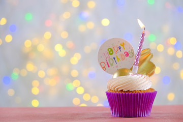 Sweet organic happy birthday cupcake with burning candle in purple wrap, cream cheese frosting swirl and golden chocolate decoration on white wooden textured table. Close up, copy space, background.