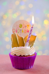 Sweet organic happy birthday cupcake with burning candle in purple wrap, cream cheese frosting swirl and golden chocolate decoration on white wooden textured table. Close up, copy space, background.