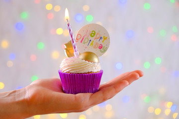 Sweet organic happy birthday cupcake with burning candle in purple wrap, cream cheese frosting swirl and golden chocolate decoration on white wooden textured table. Close up, copy space, background.