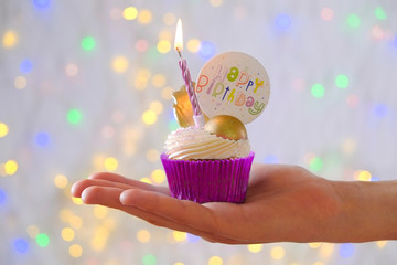 Sweet organic happy birthday cupcake with burning candle in purple wrap, cream cheese frosting swirl and golden chocolate decoration on white wooden textured table. Close up, copy space, background.