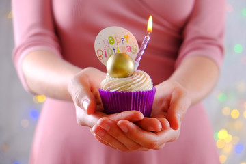 Woman holding happy birthday cupcake with burning candle in purple wrap, cream cheese frosting swirl and golden chocolate decoration on white wooden textured table. Close up, copy space, background.