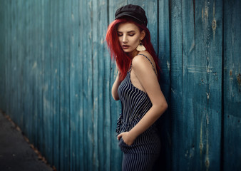 Young fashionable woman in clothes in a strip and red hair, posing on a blue background on the street.
