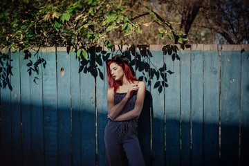 Young fashionable woman in clothes in a strip and red hair, posing on a blue background on the street.