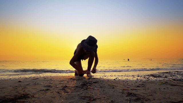Young boy with hat collecting peebles on beach and throw stone skipping game on sea sunset water surface, small flattened rock bouncing off water surface across body of water many times
