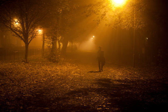 Mysterious man wearing a hat walking along an alley at night under a foggy street light