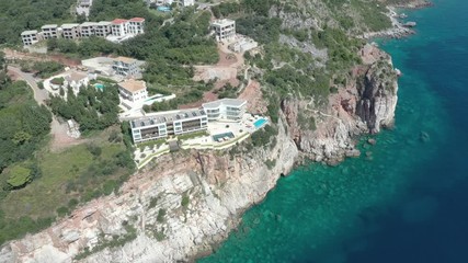Drone is flying around a steep rocky sea shore, onto which turquoise waves of the Adriatic Sea roll. On the green shores, houses are visible