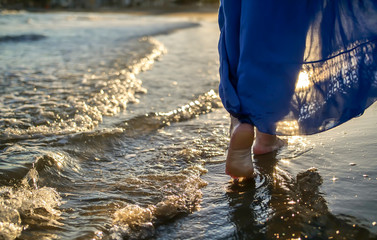 Feet of a girl in a skirt goes along the waves, glare of the sun on the water during sunset