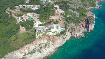 Drone is flying around a steep rocky sea shore, onto which turquoise waves of the Adriatic Sea roll. On the green shores, houses are visible