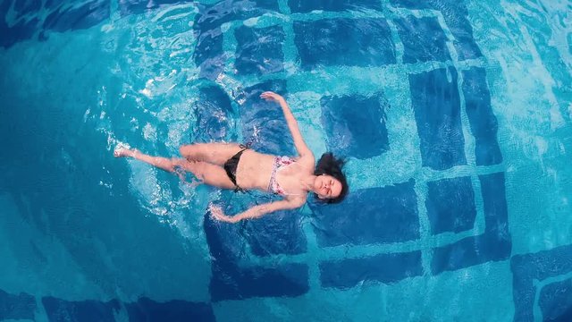 Aerial - Rotating and raising above beautiful young woman floating in crystal clear pool waters