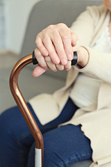 Elderly woman sitting in nursing home room holding walking quad cane with wrinked hand. Old age senior lady wearing beige cardigan, metal aid stick handle bar close up. Interior background, copy space