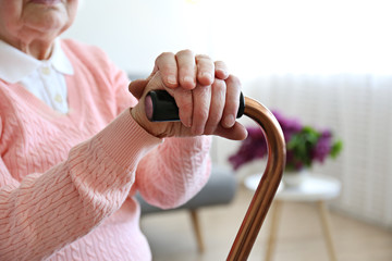 Elderly woman sitting in nursing home room holding walking quad cane with wrinked hand. Old age senior lady wearing beige cardigan, metal aid stick handle bar close up. Interior background, copy space