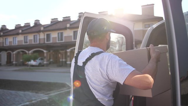 Back view of delivery man in uniform carrying cardboard box while delivering order to client living in townhouse. Adult employee of delivery service in cap and overalls taking package from car trunk