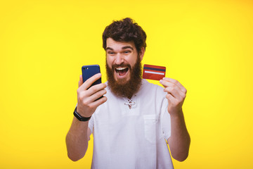 Shocked bearded guy looking at his phone while holding his red credit card on yellow background.