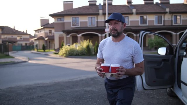 Close-up of delivery man taking coffee cups out from van and walking on road along townhouses to deliver order to home. Adult employee of food delivery service carrying coffee drinks to client