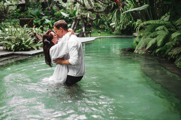 Happy couple kissing while relaxing in outdoor spa swimming pool surrounded with lush tropical greenery of Ubud, Bali. Luxury spa and wellness vacation retreat concept.