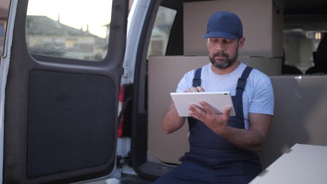 Adult delivery man in cap and overalls checking orders online on tablet sitting on edge of opened car trunk loaded with cardboard boxes. Bearded employee of delivery service networking in van