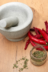 stössel und mörser auf hozbrett, daneben rote chilischoten und getrocknete kräuter im glas