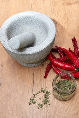 stössel und mörser auf hozbrett, daneben rote chilischoten und getrocknete kräuter im glas