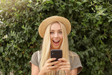 Outdoor close-up of happy young blonde woman in straw hat, keeping smartphone in hands and looking to camera joyfully, posing over green garden on sunny day