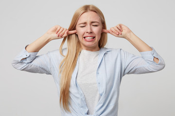 Close-up portrait of pretty young blonde woman covering her ears with hands, trying to protect herself from noise, posing over white background