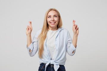 Indoor shot of a smiling young girl with long blond hair wearing blue shirt and jeans, standing over white background, holding fingers crossed for good luck and looking upwards