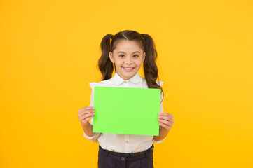 Fototapeta na wymiar Here is your homework. Adorable small child holding empty homework sheet on yellow background. Cute little girl with blank green school paper for doing homework assignment. Homework, copy space