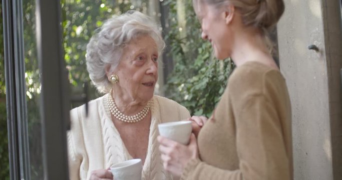 Multigeneration women talking together. Senior grandma woman smiling with her granddaughter or young friend near garden window drinking tea or coffee.White hair elderly grandmother at home.Slow motion