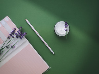 Face cream made from lavender and pink notebook with pencil on green background. Overhead shot.
