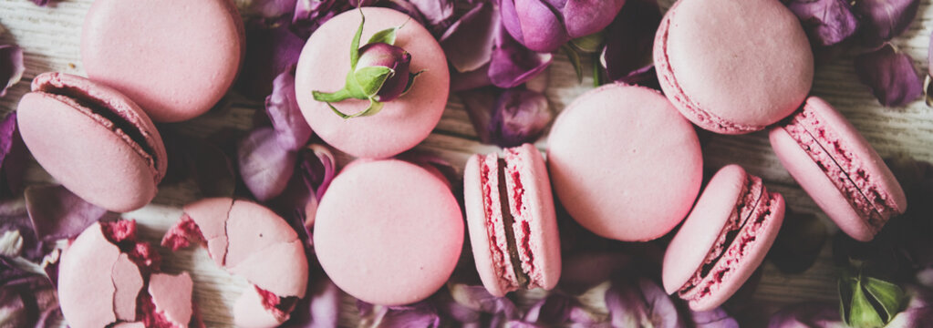 Flat-lay of sweet pink macaron cookies and rose buds and petals over wooden background, top view, close-up, wide composition. Food texture, background and wallpaper