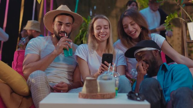 Group of millenial friends enjoying party time sitting together looking through selfie pictures on smartphone laughing of fun at summer holiday.
