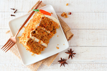 Slice of homemade carrot cake with cream cheese frosting on a plate. Top view table scene over a rustic white wood background.