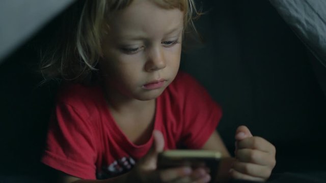 A happy boy lies in bed under a blanket and plays on a phone in a game in the dark. The face of the child is lit by a bright monitor boy under the covers Computer addict child touching phone 