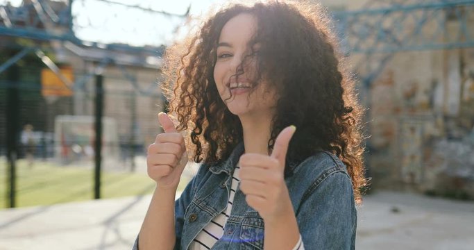Portrait shot of the cute Caucasian young pretty girl with curly hair dancing and giving her thumbs up in the slums at the courtyard on a sunny day.