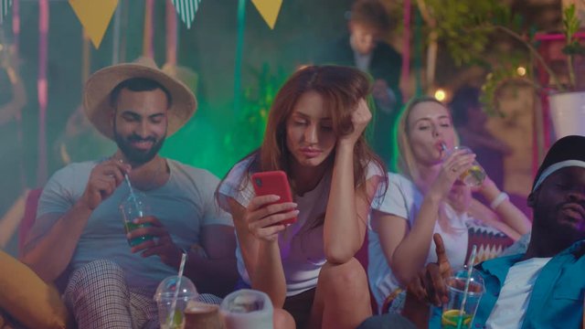 Relaxed young people chilling together in lovely summer party time. Focus on tired girl boring using mobile phone to kill time. Modern youth.