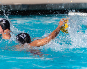 Number twelve in the black water polo uniform controls the ball while splashing and swimming toward the goal.