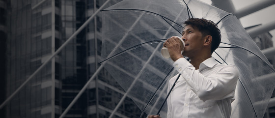 asian businessman standing and holding a cup of coffee and umbrella during rainy season in city