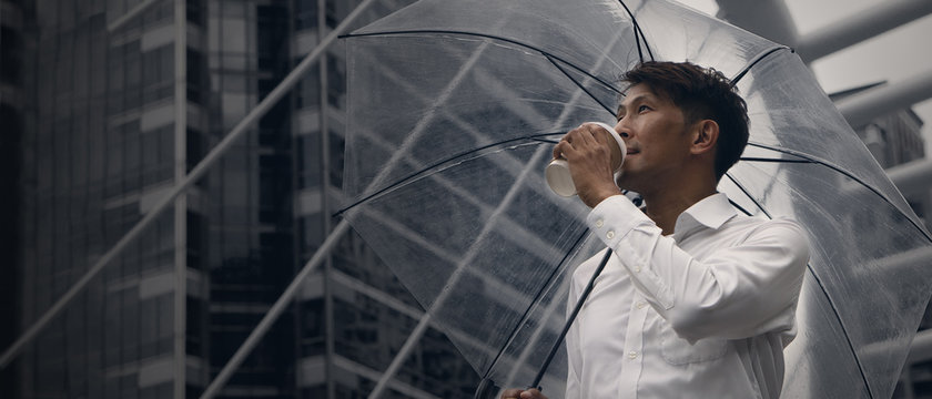 asian businessman standing and holding a cup of coffee and umbrella during rainy season in city