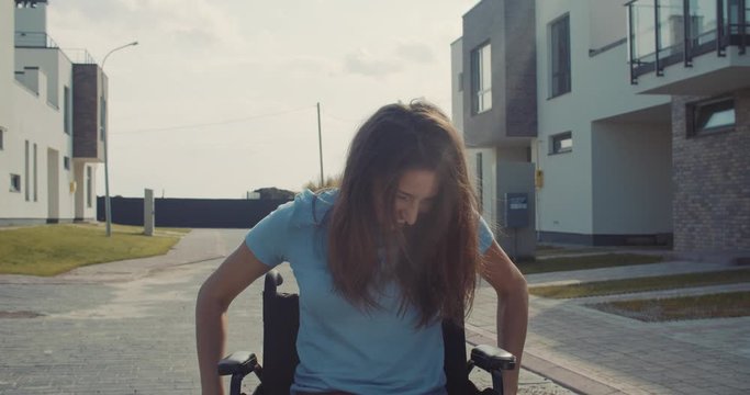 Portrait of pretty cute positive disabled sick girl trying to ride a wheelchair for the first time laughing smiling on camera staying in the street on sunny summer day.