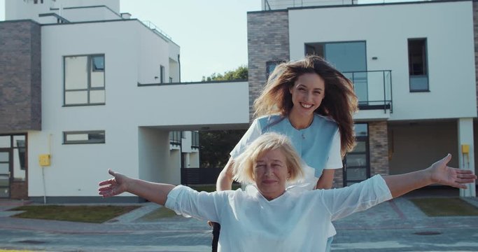 Help the elderly. Cheerful polite nurse granddaughter riding her grandpa elder disabled woman in wheelchair. Leisure fun time. Outdoors.