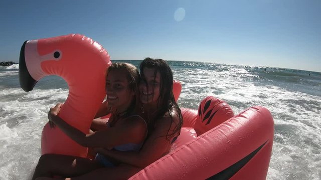 Two young women riding a blow up flamingo on ocean at the beach as waves push them around.
