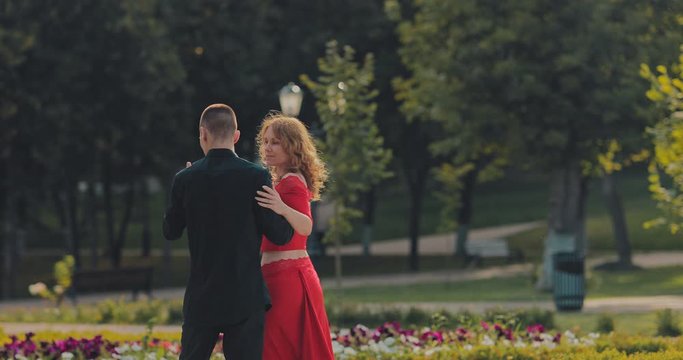 Elegant man dancing with a beautiful woman in a red tango dress in a city park. Passionate movements, body turns. Portrait view.