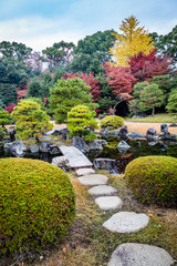 Round stepping stones lead to a stone bridge crossing water in a Japanese garden with red and yellow fall colors and round pruned shrubs