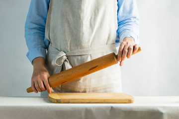 Woman cook in a cotton apron holds a wooden rolling pin at the kitchen table with a cutting board. Rustic style.