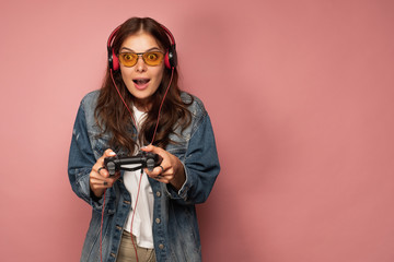 Brunette on a pink background in jeans, glasses and headphones in amazement looking forward,...