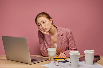 Red-haired girl in a jacket sitting at a table in front of a laptop on a pink background, his head on his hand, falling asleep.